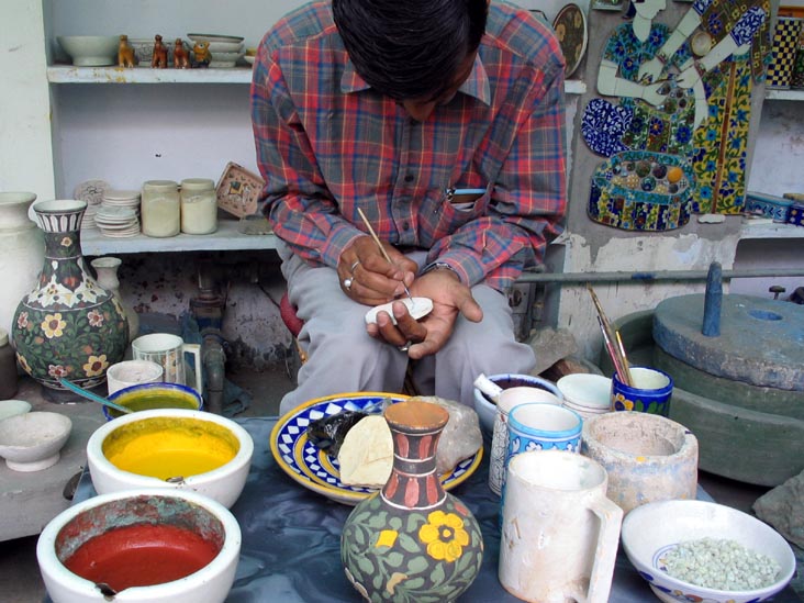 Craftsman using a fine brush to paint a small pottery piece at the Jaipur Blue Pottery Art Centre, surrounded by bowls of colour.